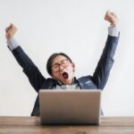Yawning - Photo of Yawning Man with His Hands Up and Eyes Closed Sitting at a Table with His Laptop